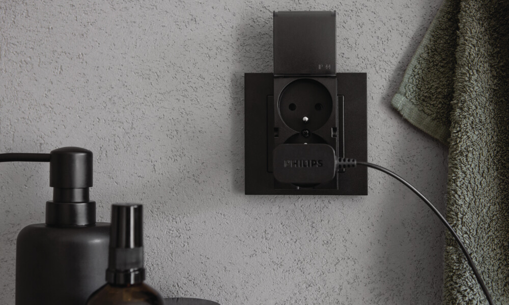 Czarne podwójne, bryzgoszczelne gniazdo elektryczne do łazienki z matową klapką z serii Simon 55 zamontowane na ścianie.
